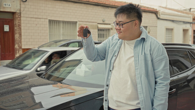 Young man holding car key up and standing by a parked car on a residential street, smiling and checking key function; quiet pride.