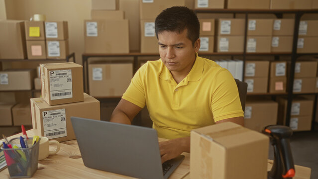Man typing on laptop among cardboard boxes with hands on keyboard in a building warehouse shipping area; frustration small business.
