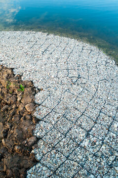 Geocell shore reinforcement background. A pebble filled cellular grid covering a lake bank. Dark soil and clear water visible at the edge of the armored shore