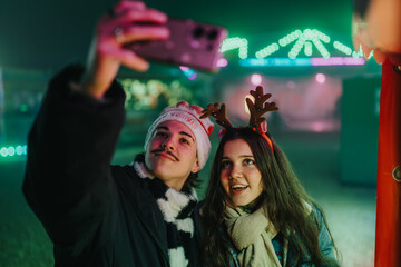 Naklejka premium Two friends wearing holiday accessories take a selfie at a nighttime fair. A young man in a Santa hat and a woman with reindeer antlers smile under colorful lights.