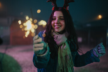 Naklejka premium A young woman wearing reindeer antlers smiles while holding a lit sparkler. The festive night scene shows a joyful friend celebrating outdoors with bokeh lights.