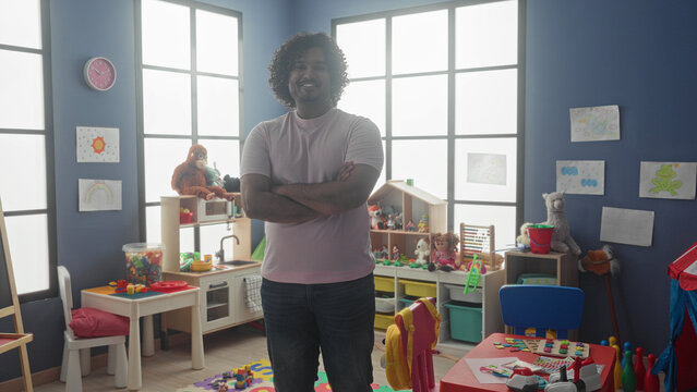 Man arms crossed standing amid toys and cubbies in a kindergarten building, young teacher smiling at camera; nurturing calm learning.
