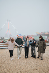 Naklejka premium A group of five friends walking and laughing at a winter amusement park while wearing Santa hats and warm coats. The friends enjoy a festive outdoor holiday outing near rides and colorful stalls.
