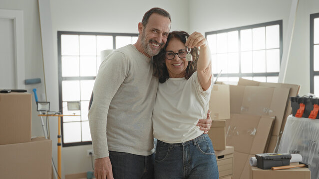 Man and woman embrace as she holds a house key aloft beside stacked moving boxes inside a building; new home joy.