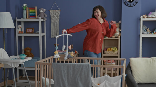 Woman touching mobile above wooden crib while dancing and smiling inside a home building nursery room; tender nurturing joy.