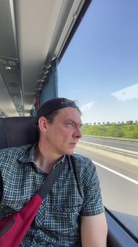 seated male passenger in plaid shirt and backward cap looks through large coach window at blurred fields and roadside greenery under clear blue sky sunlight man