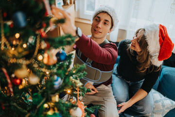 Naklejka premium A young couple decorates a Christmas tree at home while wearing Santa hats. They share a warm holiday moment arranging ornaments and lights together.