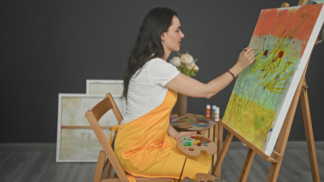 Woman painter in studio holds brush and palette with hand while applying paint to canvas and smiling; creative joyful.