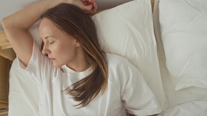 Woman lying in bed with arm above head and hand on stomach on white sheets beside a rattan nightstand in building; serenity rest.