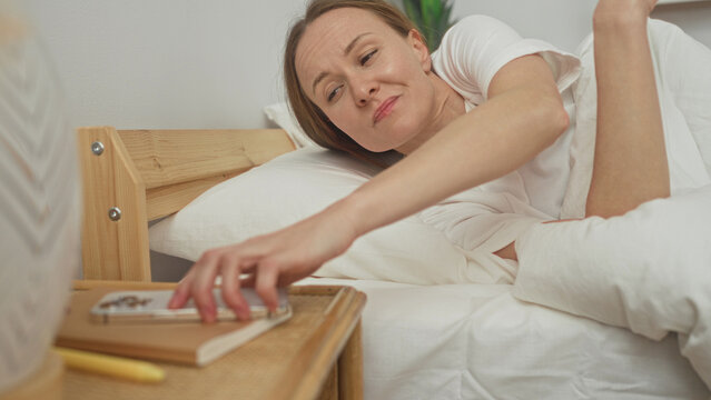 Woman reaches for smartphone on bedside table, hand outstretched while half lying under white duvet in a bedroom inside a residential building; morning frustration.