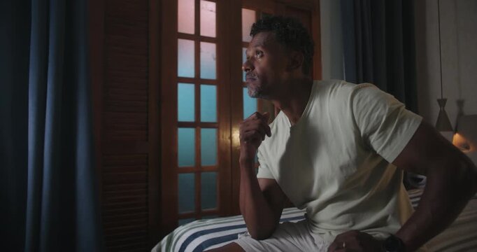 Man of African descent sitting on bed, resting hand on chin, looking away in deep contemplation, introspective mood, serious expression, quiet indoor setting
