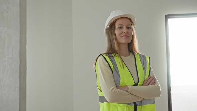 Woman in hardhat and hi vis vest standing with arms crossed inside building under construction, concrete column visible; confidence leadership.