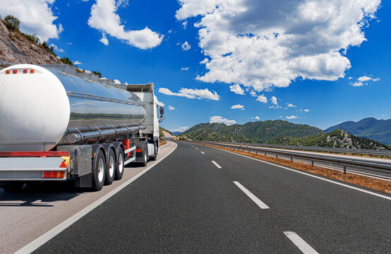 A white tanker truck drives along a scenic mountain highway under a clear blue sky with fluffy white clouds.