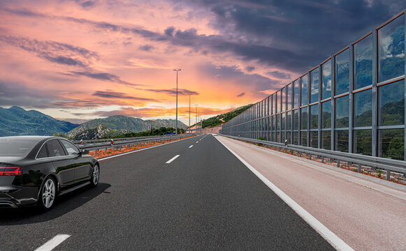 A modern black sedan drives along an empty highway with a noise barrier against a scenic mountain backdrop under a colorful sunset sky.