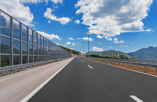 An empty modern highway stretches through a mountainous landscape under a bright blue sky with fluffy white clouds.