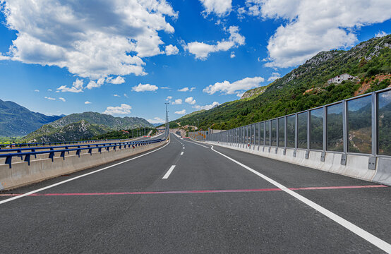 An empty modern highway stretches through a scenic mountain landscape under a bright blue sky filled with white clouds.