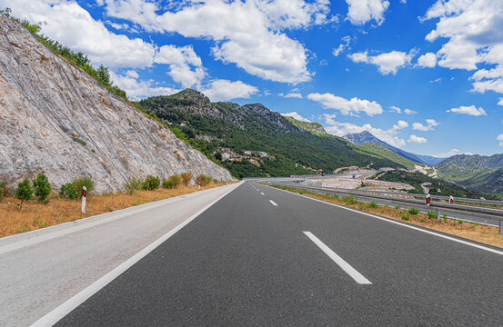 An empty asphalt road stretches through a scenic mountain landscape under a bright blue sky filled with white clouds.