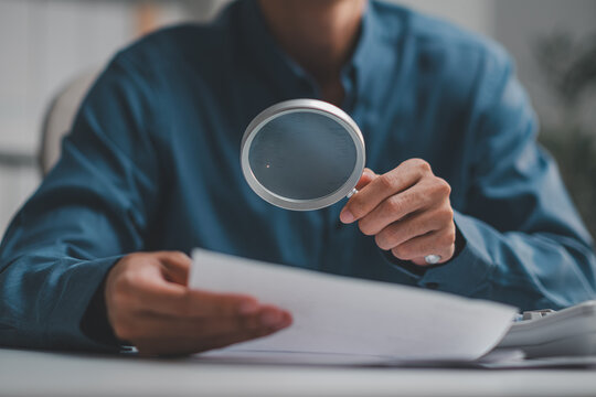 Businessman holding magnifying glass to inspect and examine contract documents for corporate audit and risk analysis