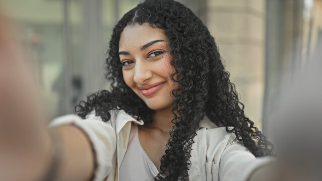 Young arab woman smiling selfie with visible face and arm extended on city street near glass windows; confidence.