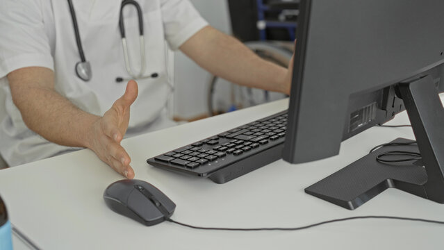 Man doctor with stethoscope reaching for computer mouse and typing on keyboard at desk in building; focused care.