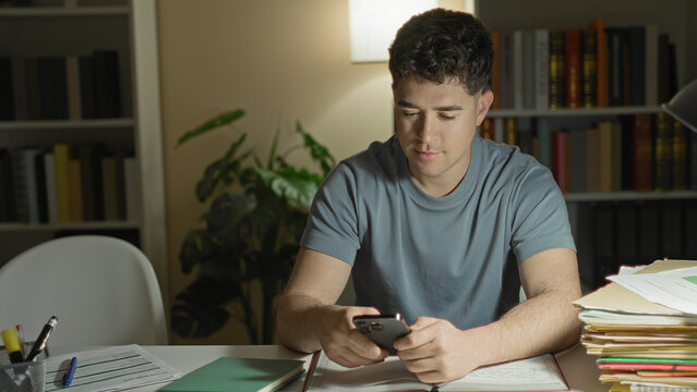 Young man holding smartphone with both hands, looking down at papers and a stack of folders on a desk in a building; quiet concentration.