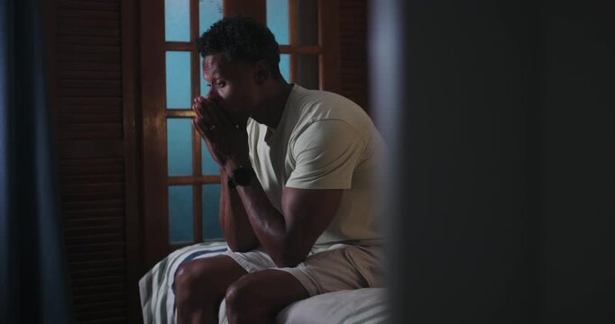 One Black man sitting on bed with hands covering face, overwhelmed emotion, deep contemplation, serious mood, introspective moment in quiet indoor setting with wooden shutters