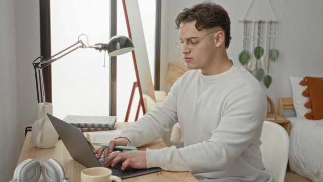 Man typing on laptop and jotting notes with pen, hand on keyboard at wood desk beside lamp and book in bedroom; concentration study focus.