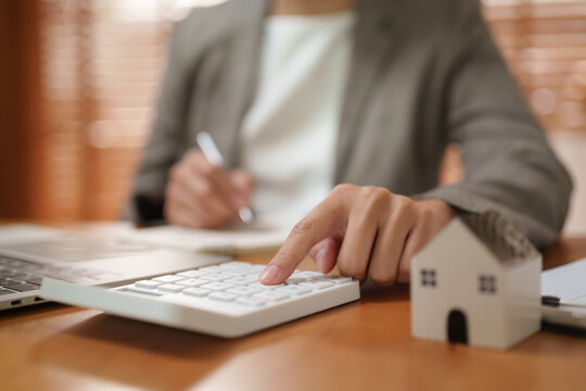 Businessoman using a calculator beside a miniature house while reviewing finances at a desk, representing mortgage planning, home loans, property costs, budgeting and real estate investment.