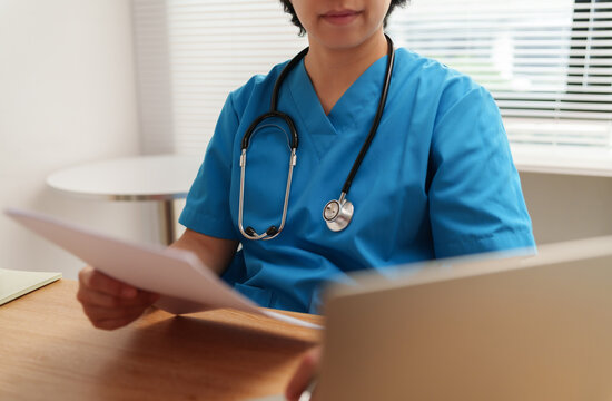 Asian Healthcare professional in blue scrubs reviewing medical documents beside a laptop, representing patient care, diagnosis, hospital work and digital healthcare.