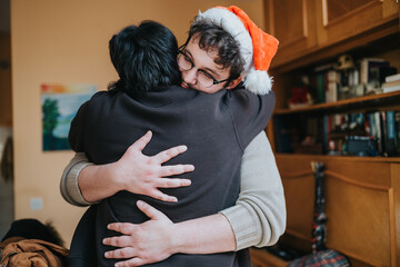 Naklejka premium Two people embrace indoors while one wears a Santa hat. The warm holiday hug captures friendship and festive togetherness at home.
