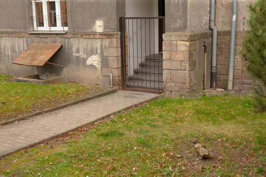 Grassy approach path leading to gated stair and stoop, worn pavement edging with scattered leaves and moss, small front yard and muted tones conveying peaceful residential arrival 