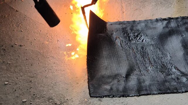 Construction worker installing bituminous waterproofing membrane on a flat roof, heating the material with an open flame torch and smoothing the edges by hand.
