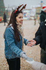 Naklejka premium A young woman in a denim jacket and scarf wearing reindeer antlers smiles while reaching for hands. She enjoys a festive moment at an outdoor Christmas market with people holding hands.