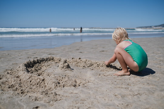 girl in green swimsuit sitting on sandy beach building sandcastles by ocean