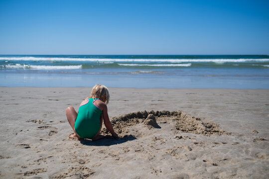 girl in green swimsuit sitting on sandy beach building sandcastles by ocean