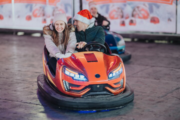 Obraz premium A young couple rides together in a bright orange bumper car, smiling and laughing. They enjoy a festive amusement park ride at a winter fair with holiday decorations.