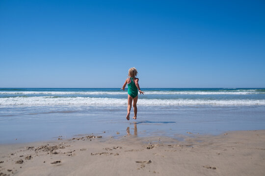  little girl jumping on wet sand beach into ocean waves on sunny summer day