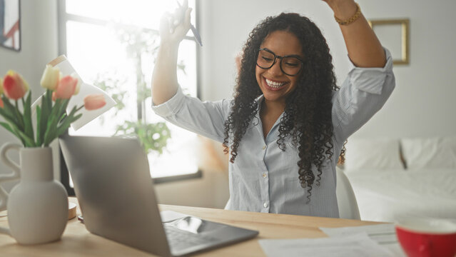 Woman at laptop raises arms and claps smiling in a home office studio with tulips and papers on desk; work success celebration.