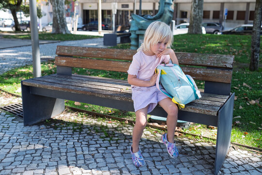 Sad little girl sitting alone on park bench with backpack