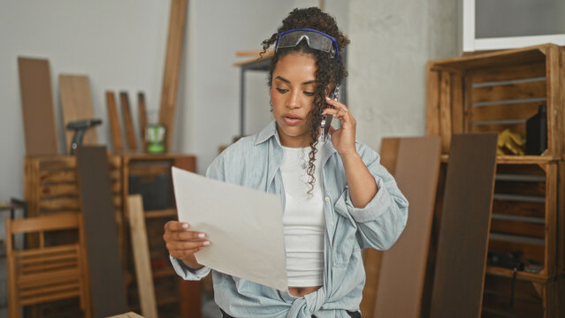 Woman carpenter holding smartphone to ear and reading a sheet of plans in studio workshop amid wooden planks and crates; concentration planning.