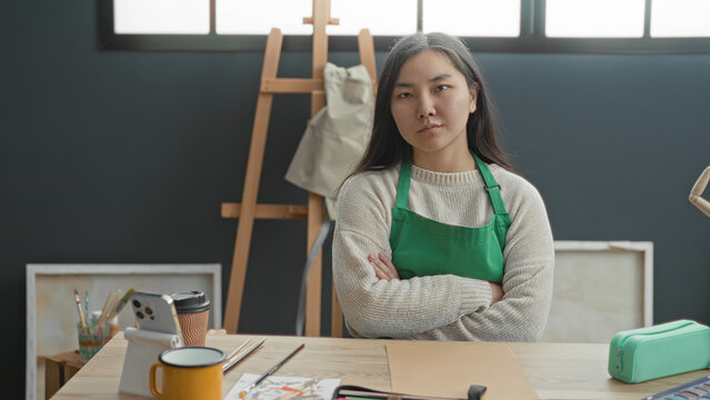 Woman wearing green apron with arms crossed at studio beside easel and painting tools on table, neutral expression and seated posture; determination focus.