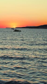 Sunset casts vibrant orange and yellow colors over the ocean, with boats gently bobbing on the water's surface, creating a serene evening atmosphere