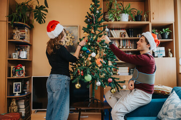 Naklejka premium A young couple decorates a Christmas tree in their living room while wearing Santa hats. They adjust ornaments and lights on the tree, creating a cozy holiday scene.