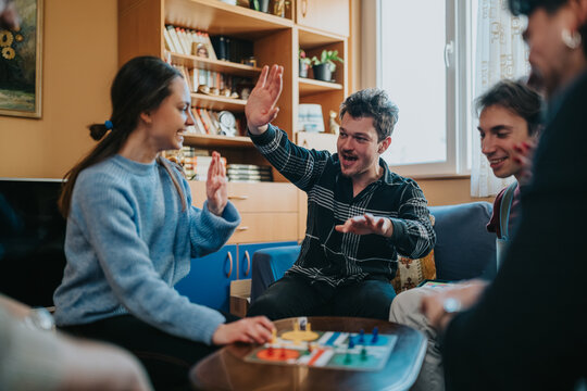 A group of friends play a colorful board game together in a cozy living room. They laugh, high-five and enjoy a relaxed game night at home.
