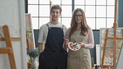 Woman painter holding palette and brush, smiling beside easel and man wearing apron in studio  creativity collaboration joy. © Krakenimages.com