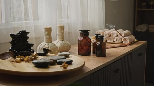 Incense stick emitting rising smoke beside massage stones, herbal compress balls, and essential oil bottles in wellness center. Footage ideal for spa salon advertising and relaxation therapy promo