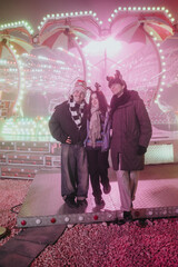 Naklejka premium Three friends dressed in winter clothes smile together at a brightly lit carnival ride. The festive scene shows holiday accessories and colorful lights at a nighttime amusement park.