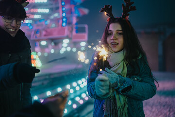 Naklejka premium Two friends hold sparklers while enjoying a festive winter night. The scene shows a young pair celebrating among colorful lights at a holiday market.