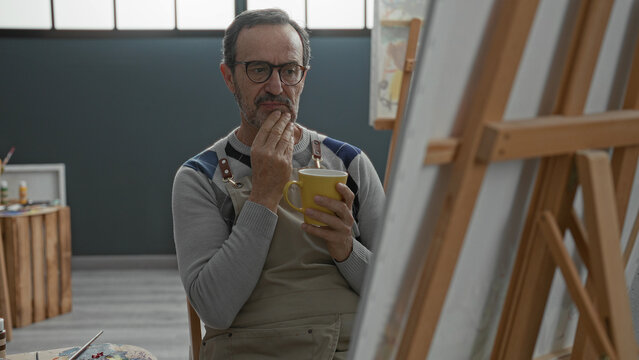 Man painter in apron holding yellow mug with hands, gazing at canvas on easel in studio; contemplative solitude.