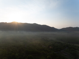 Aerial sunrise over misty tropical forest and mountain ridge in rural Thailand, warm sun rays illuminating valley at dawn with greenery, morning fog. Nature, travel, tranquility and environment themes © Fahroni